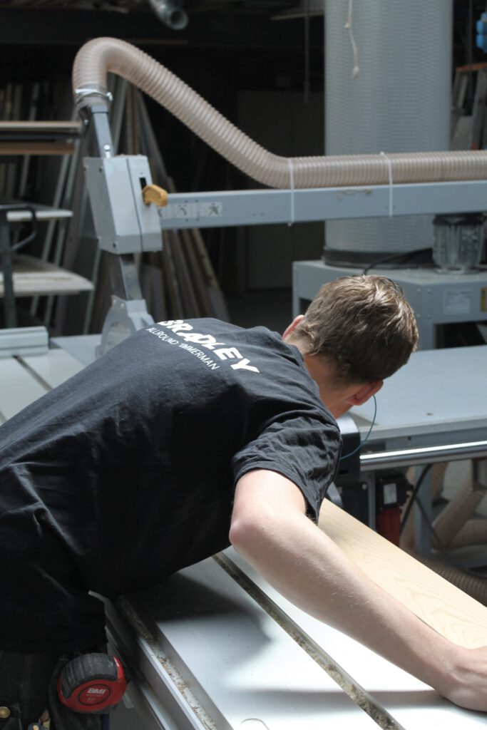 Bradley, allround timmerman, werkt met uiterste precisie aan een stuk hout in zijn werkplaats. Dankzij professioneel gereedschap en vakkennis levert hij maatwerk van topkwaliteit.